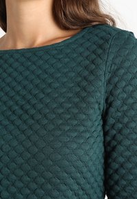 Suéter texturizado en tono verde azulado, con un patrón de diamantes, cuello redondo y tejido texturizado; acabado suave con un aspecto estructurado.