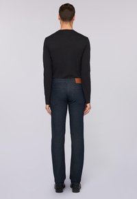 Jeans de un azul oscuro con un corte recto, que cuentan con bolsillos traseros y una etiqueta de cuero marrón en la cintura. Combinados con una camiseta negra de manga larga.