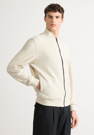 Ung man i en ljusbeige zip-up jacka med hög krage och svarta byxor, poserande med en hand i fickan mot en enkel bakgrund.