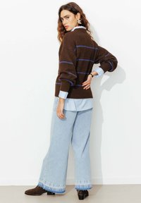 Pull marron tricoté avec des rayures bleues, superposé sur une chemise bleu clair. Pantalon en denim bleu clair à jambes larges avec ourlets effilochés et bottes marron.