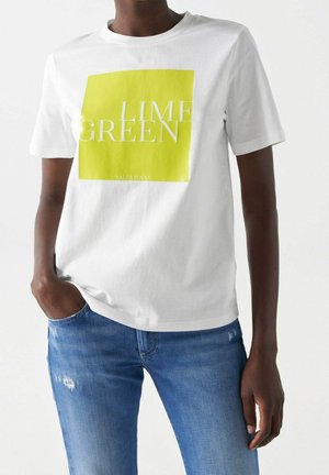 Persoon draagt een wit T-shirt met een geel vierkant en de tekst ‘LIME GREEN’, gecombineerd met een blauwe spijkerbroek, één hand in de zak.