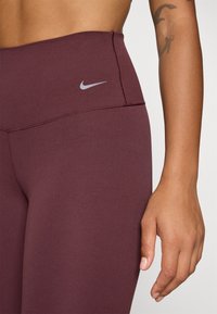 Bordo tajice viskog struka s glatkom teksturom, koje imaju sivi Nike logo na pojasu i ojačane detalje šivanja.