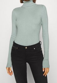 Pull en maille côtelée vert clair à col roulé avec des manches longues, doté de trous pour les pouces aux poignets, associé à un jean noir et des accents dorés.