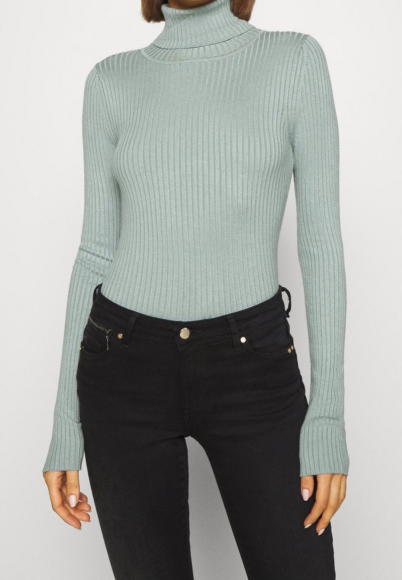Pull en maille côtelée vert clair à col roulé avec des manches longues, doté de trous pour les pouces aux poignets, associé à un jean noir et des accents dorés.