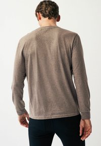 Langärmliges Hemd in Taupe, strukturierter Stoff, mit abgerundetem Saum und glattem Rücken; kombiniert mit dunklen Hosen.
