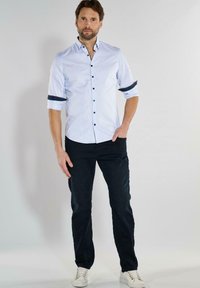 Himmelblau gestreifte Button-Up-Bluse mit dunklen Knöpfen und hochgekrempelten Ärmeln, kombiniert mit dunklen Hosen und weißen Sneakers.