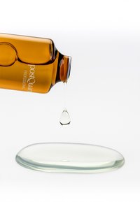 Botella ámbar con etiqueta "PostQuam Professional" vertiendo una gota de líquido claro sobre una superficie lisa y transparente.