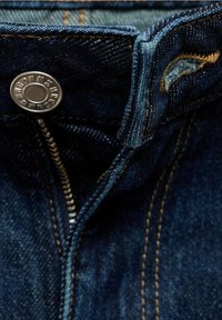 Gros plan d'un jean en denim bleu foncé avec une fermeture éclair ouverte et un bouton en métal marqué "MNG."
