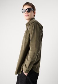 Olive green, long-sleeve shirt made of soft fabric with a button-down collar and single chest pocket, paired with large, angular sunglasses.