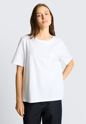 Młoda kobieta z prostymi brązowymi włosami, ubrana w prostą białą koszulkę z kieszonką i ciemnoniebieskie dżinsy, stojąca na jasnym tle.