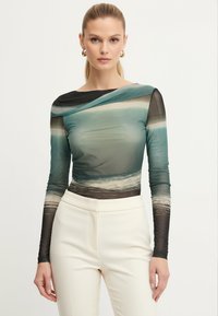 Top in rete a maniche lunghe, caratterizzato da una sfumatura di teal e nero con un motivo a onde, scollatura rotonda e vestibilità slim. Abbinato a pantaloni bianchi.