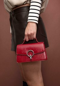 Red leather handbag with a structured shape, featuring a flap closure, a circular metal accent, and a top handle. Textured finish.