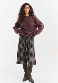 Bordowy miękki sweter z szerokimi rękawami, zestawiony z wzorzystą brązową spódnicą midi w kratę i brązowymi kozakami za kolano. Prosty akcent w postaci naszyjnika.
