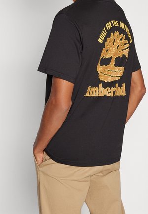 Personne portant un t-shirt Timberland noir avec un logo arbre jaune et le texte "Built for the outdoors" au dos, associé à un pantalon beige.