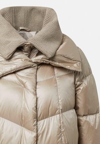 Beige pufferjack met een getextureerde gebreide kraag. Het ontwerp heeft een chevron-quiltpatroon en een glanzende afwerking.