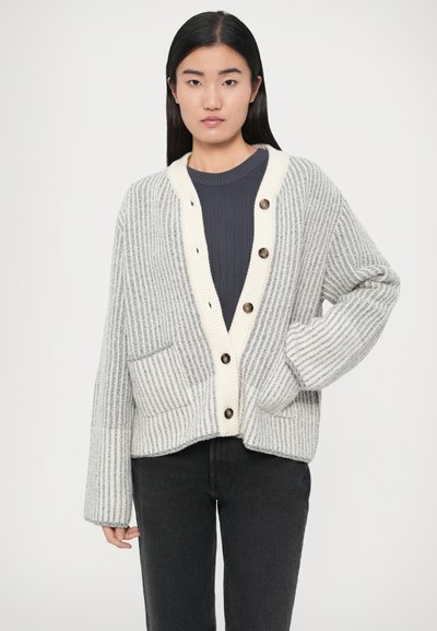 Καρδίγκαν με γκρι και λευκές ρίγες, V-λαιμόκοψη, κρεμ γιακά, πέντε καφέ κουμπιά και δύο εμπρόσθιες τσέπες. Μαλακή πλεκτή υφή.