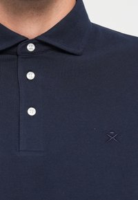 Közelről látható egy személy, aki sötétkék polo inget visel, három fehér gombbal és egy kisméretű hímzett logóval a mellkasán.