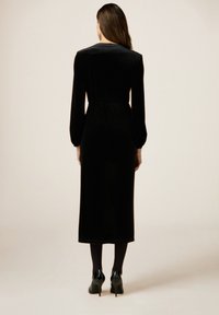 Abito in velluto nero con maniche lunghe, vita adattata e lunghezza midi. Texture liscia, ornamenti minimi, indossato con tacchi neri.