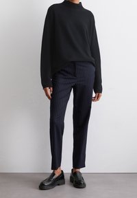Pull noir en maille avec col montant, associé à un pantalon bleu marine rayé. Chaussures : mocassins noirs à semelle épaisse.