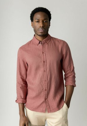 Man staat tegen een effen achtergrond, gekleed in een lichtroze overhemd met lange mouwen die zijn opgerold en lichtbeige broek.