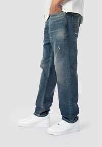 Moški stoji z rokami v žepih, oblečen v zbledela modra jeans hlače in bele superge, pred enobarvno ozadjem.