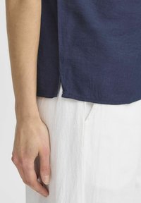 Top in lino blu navy con maniche corte, tessuto testurizzato e spacchi laterali, abbinato a pantaloni bianchi testurizzati. Focus su linee pulite e semplicità.