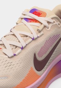 Chaussure de course Nike beige avec des lacets blancs, des accents violets et orange, un swoosh marron foncé, et le texte "RUN" sur l'étiquette de la languette.