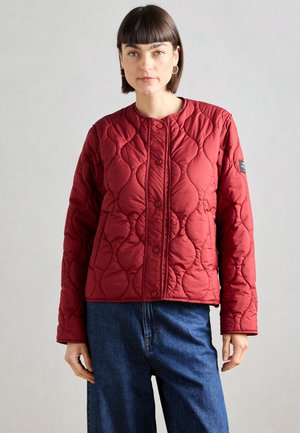 Red quilted jacket with a rounded collar and snap button closure. Features a textured pattern and long sleeves. Worn with blue jeans.