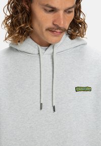 Sudadera con capucha gris de algodón suave, que presenta un parche de logo texturizado en azul y amarillo en el lado izquierdo del pecho, con detalles de cordón.