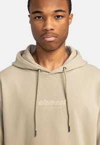 Beige hættejakke med snore, som har en blød tekstur og et subtilt logo tryk med teksten "element skateboard co." på forsiden.