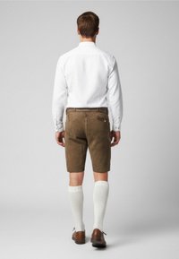 Pantaloni corti in suede marrone con tasche, abbinati a calze bianche fino al ginocchio e scarpe marroni, indossati sotto una camicia bianca a maniche lunghe.