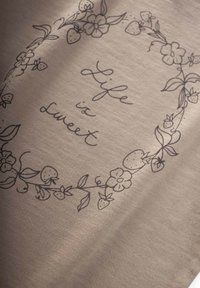 Beige Stoff mit schwarzer Blumen- und Erdbeerillustration, zeigt den Text "Das Leben ist süß" in einer fließenden Schrift, die ein einfaches Design vermittelt.