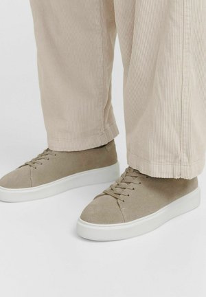 Baskets en daim beige avec des lacets plats et une semelle en caoutchouc blanche. Portées avec un pantalon en velours côtelé beige clair, montrant une coupe décontractée et une texture.