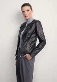 Kobieta ubrana w czarną skórzaną kurtkę na szarym golfie i szare spodnie w prążki, stojąca bokiem z jedną ręką w kieszeni.