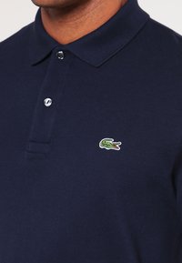 Mornarsko modra polo majica iz teksturirane tkanine; ima tri gumbe na zapenjanju in zeleni vezeni krokodilov logotip na prsih.