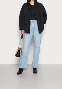 Persona con una chaqueta de plumas negra, parte superior negra, jeans rectos de mezclilla azul claro, botas negras y sosteniendo un bolso bicolor con cadenas doradas.