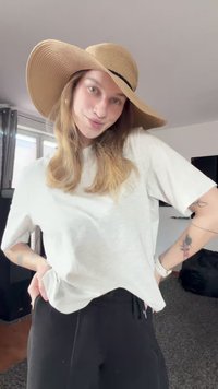 Mlada osoba s dugom kosom, koja nosi veliki sunčani šešir, bijelu majicu i crne hlače, pozira unutra s rukama na bokovima.