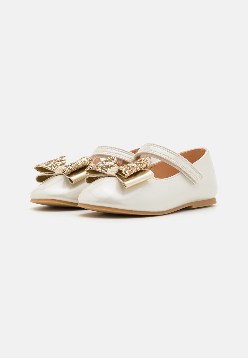 Weiße Lacklederschuhe mit flacher Sohle, ausgestattet mit einer großen, glitzernden goldenen Schleife und zwei elastischen Riemen über dem Spann.