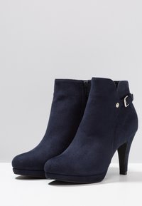Botas de tobillo de ante azul marino con punta afilada, cremallera lateral y un detalle decorativo de hebilla. El tacón de aguja añade altura.