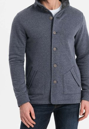 Mand iført en knappet, mørkegrå jakke med to forlommer og blå jeans, står mod en ensfarvet hvid baggrund.