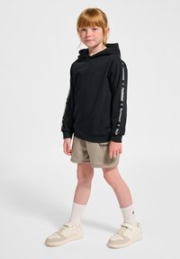 Schwarze Kapuzenjacke mit Logo auf den Ärmeln, kombiniert mit beigen Shorts und hellen Sneakern. Das Model hat rote Haare und trägt weiße Socken.