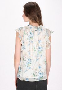 Bluse mit floralem Muster aus leichtem, transparentem Stoff, mit Rüschensaum an Kragen und Ärmeln. Die Farben umfassen blaue, grüne und cremefarbene Blumenmuster.