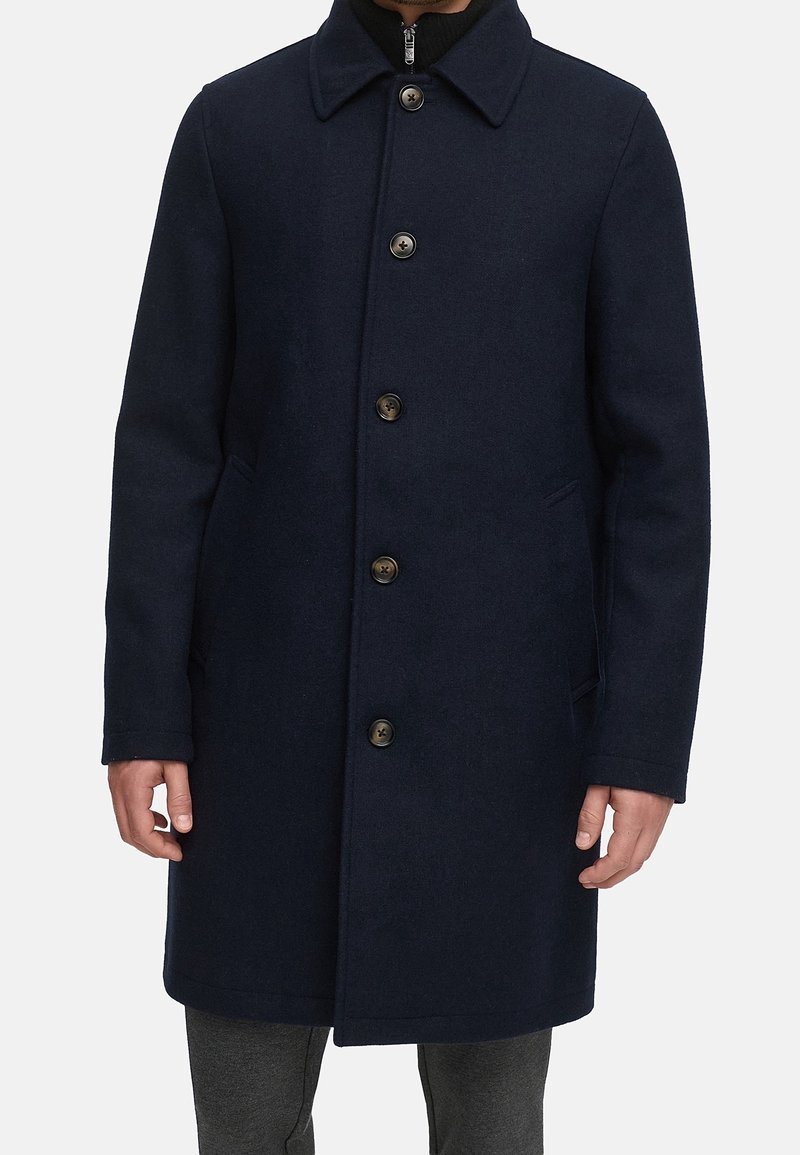 Homme portant un manteau en laine bleu marine foncé à simple boutonnage, avec des poches latérales et un col, par-dessus un haut noir zippé.