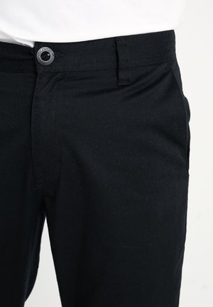 Czarne spodnie z zapięciem na guzik i przednimi kieszeniami, noszone z włożoną białą koszulą, ukazujące talię i górną część uda.