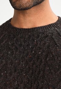 Nahaufnahme des unteren Gesichts und des Halses eines Mannes, der einen dunkelbraunen, strukturierten Zopfmuster-Pullover mit einem gerippten Rundhalsausschnitt trägt.