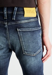 Jeans in denim blu scuro con una texture sbiadita. Include una toppa in pelle gialla con la scritta "REPLAY." Presenta tasca posteriore con dettagli cuciti.