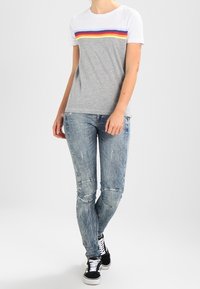Camiseta de manga corta en gris y blanco con rayas horizontales en rojo, amarillo y azul, combinada con jeans azules desgastados y zapatillas negras.