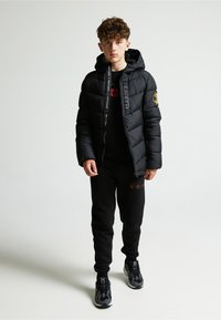 Melna pūkainā jaka ar kapuci, ar rāvējslēdzēja aizdari un logotipa uzšuvumu. Valkāta virs melnas krekla un melnām sporta biksēm, papildināta ar melniem sporta apaviem.