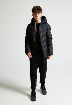 Chaqueta de plumas negra con capucha, cierre de cremallera y parche con logo. Llevada sobre una camiseta negra y pantalones de chándal negros, complementada con zapatillas negras.