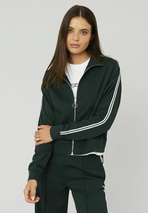 Femme aux longs cheveux bruns portant une veste de survêtement verte foncée et un pantalon avec des rayures blanches, se tenant contre un fond clair.